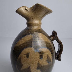 Może przedstawiać: Ceramiczny dzbanek o pękatym korpusie i rozszerzanym, falistym rancie. Naczynie pokryte jest ciemnobrązowym szkliwem z jasnobrązowymi, abstrakcyjnymi, wirującymi wzorami oraz subtelnymi niebieskimi akcentami. Posiada jeden zakrzywiony uchwyt z boku.