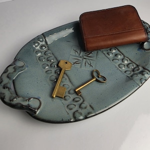 May include: An oval-shaped, blue-grey ceramic tray with textured, raised circular patterns and two side handles. Resting on the tray are a brown leather wallet and two brass-colored metal keys, one with a standard head and one with a heart-shaped bow.