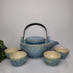 May include: A ceramic tea set featuring a teapot with a black handle and a matching lid, accompanied by four small cups. The pieces are finished in a glossy blue glaze with a textured, embossed floral pattern and cream-coloured interiors.