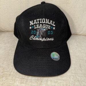 Puede incluir: Una gorra de béisbol negra con bordados en blanco y azul cerceta que dicen National League 2003 Champions, un logotipo F estilizado y un gráfico de un marlín. Una pegatina holográfica de autenticación de la MLB está pegada en la visera.