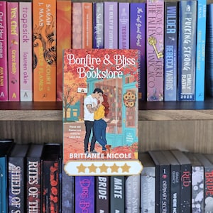 May include: A paperback book titled Bonfire & Bliss Bookstore by Brittanee Nicole, featuring an illustration of two people embracing in front of a shop. The book is displayed on a shelf in front of other books, with a five-star rating graphic below it.