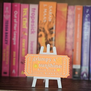 May include: A rectangular orange ticket-shaped sign on a small white easel. The text reads "grumpy x sunshine BOOK CLUB LIFETIME MEMBERSHIP" with a sun icon, a cloud icon, and two small hearts. The ticket includes "ADMIT ONE" and "02631224".