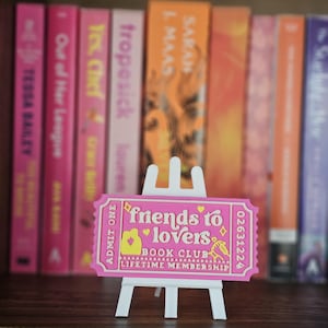 May include: A pink rectangular ticket-shaped sign on a white easel stand. The sign features the text "friends to lovers BOOK CLUB LIFETIME MEMBERSHIP" and "ADMIT ONE" with yellow graphic icons of a couple, hearts, and a small book illustration.