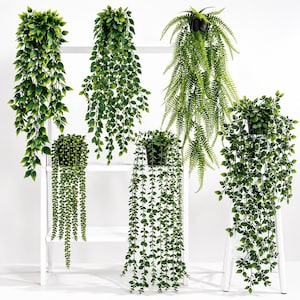 May include: A collection of six artificial hanging plants in black pots, including a string of pearls, a fern, and various trailing leafy vines with green foliage, displayed on a white tiered shelving unit against a plain white background.