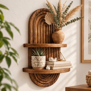 May include: A wall-mounted wooden shelf unit featuring a dark brown finish with a vertical arched back panel. The unit has two horizontal shelves holding a terracotta vase with dried foliage, a patterned succulent pot, books, and a gold pyramid.