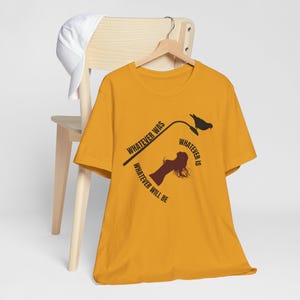 Może przedstawiać: Musztardowy t-shirt z krótkim rękawem z czarną grafiką przedstawiającą ptaka na latarni ulicznej oraz sylwetkę osoby. Tekst brzmi: WHATEVER WAS, WHATEVER IS, WHATEVER WILL BE. Biała czapka z daszkiem wisi na drewnianym wieszaku nad koszulką.