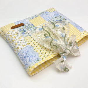 May include: A quilted fabric cover featuring a patchwork design of yellow gingham, blue floral patterns, and small yellow flowers. A sheer white lace overlay with green and blue embroidered floral vines is attached to the front with a brown tag.