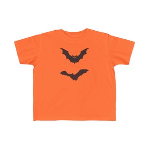 May include: A bright orange short-sleeve t-shirt featuring two dark brown bat silhouettes printed on the centre chest. The top bat has two small yellow eyes, while the bottom bat is a solid dark brown silhouette with wings spread wide.