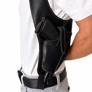 Könnte beinhalten: Ein schwarzes Schulterholster aus Leder mit weißen Kontrastnähten, einem sicheren Riemenverschluss und einer geformten Tasche für eine Handfeuerwaffe. Das Holster ist an einem breiten Schultergurt befestigt und mit einem schwarzen Gürtel verbunden.