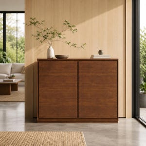 May include: A rectangular wooden cabinet with two flat-panel doors featuring a rich brown finish. On top sits a white ceramic vase containing a leafy green branch, a small dark bowl, two stacked white books, and a small grey spherical object.