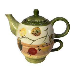 May include: A ceramic tea for one set featuring a teapot stacked on a matching teacup. The set is cream-colored with hand-painted fruit motifs including yellow and red fruits, green leaves, and a green rim with white cross-stitch detailing.