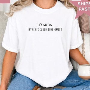 May include: A white short-sleeved t-shirt featuring the black printed text IT'S GIVING HYPERFOCUSED SIDE QUEST centered on the chest. The garment has a crew neckline and a relaxed fit, paired with blue denim jeans and a gold chain necklace.