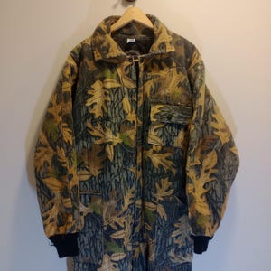 May include: A full-body jumpsuit featuring a realistic forest camouflage pattern with brown, tan, and green oak leaf motifs. The garment includes a collared neckline, a front button closure, a chest pocket with a flap, and black ribbed cuffs.