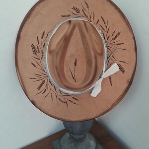 Op de afbeelding: Een beige hoed met brede rand, voorzien van donkerbruine, met de hand geschilderde tarwestengels op de rand en de bol. De hoed heeft een gerafelde lichtblauwe stoffen band rond de basis, geaccentueerd met een kleine witte stoffen strik.