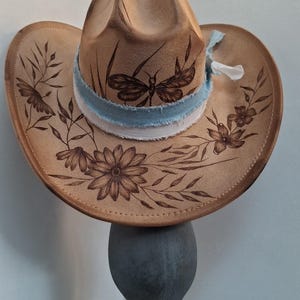 Op de afbeelding: Een beige cowboyhoed met donkerbruine pyrografie-illustraties van een libel en diverse wilde bloemen. De hoed is voorzien van een gelaagde, gerafelde lichtblauwe en witte stoffen band die rond de basis van de kroon is gewikkeld.