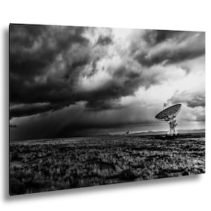 May include: A black and white photograph of a large satellite dish antenna standing in a vast, flat field of dry grass under a dramatic, dark, and heavy storm cloud sky. The scene captures the contrast between the mechanical structure and nature.