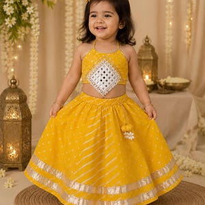 May include: A bright yellow two-piece outfit consisting of a halter-neck crop top and a matching flared skirt. The top features a central diamond-shaped mirror embellishment. The skirt has white diagonal stripes, polka dots, and silver trim.