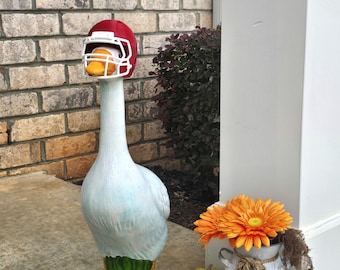 May include: A white lawn goose statue wearing a red football helmet with a white face mask. The goose stands on a green base. Next to it is a galvanized metal milk can containing orange flowers, a small gold pumpkin, and a white rock.