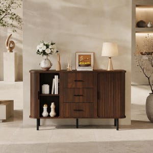 May include: A dark wood sideboard featuring three central drawers with black horizontal handles, flanked by two side cabinets with vertical fluted doors. The unit stands on four black legs and holds a vase of white flowers, a lamp, and decor.