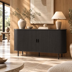 May include: A black sideboard with a fluted, rounded-edge design and two central doors. It stands on thin black legs and holds a textured beige vase with greenery, a small bowl, a stack of books, and a table lamp with a beige fabric shade.