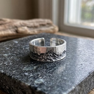 May include: A silver-colored open band ring featuring a textured, dark, rugged landscape relief pattern along the lower half and a smooth, polished finish on the upper portion, creating a distinct contrast in surface depth and metallic sheen.
