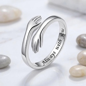 May include: A silver-toned metal ring featuring a design of two hands wrapping around the band in a hugging motion. The outer surface of the band is engraved with the black text Always with you in a cursive script font, creating a continuous loop.