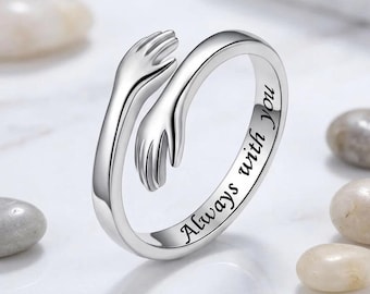 May include: A silver-toned metal ring featuring a design of two hands wrapping around the band in a hugging motion. The outer surface of the band is engraved with the black text Always with you in a cursive script font, creating a continuous loop.
