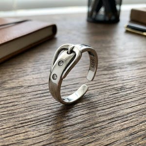 May include: A silver-colored open ring shaped like a belt buckle, featuring two small clear stones embedded in the front section. The interior of the band is clearly stamped with the text S925, indicating the metal composition of the jewelry piece.