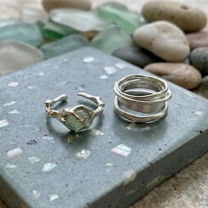 May include: Two silver rings. One ring features an open, textured band with a central pale green stone. The second ring consists of multiple stacked, smooth silver bands that form a single cohesive piece with a rounded, layered appearance.