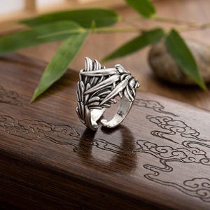 May include: A silver-toned metal ring featuring an intricate design of layered, textured feathers that wrap around the band. The ring has an open-back construction, allowing for adjustable sizing, and displays a polished finish with darkened recessed areas.