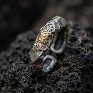 May include: A silver-toned metal ring with an open band design featuring a hammered, textured surface. The center of the ring displays a raised, gold-colored sun motif with intricate swirling patterns and radiating rays engraved into the metal.