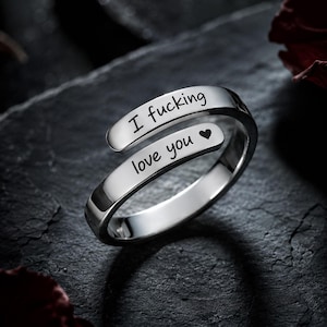 May include: A polished silver-tone bypass ring featuring an open design. The top band is engraved with the text I fucking and the bottom band is engraved with the text love you followed by a small heart symbol in a clean black script font.