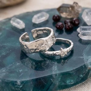 May include: Two silver rings with an open band design, each stamped with S925. The wider ring features a hammered texture with an engraved star, while the thinner ring has a prominent star-shaped accent. Both rings have a polished finish.