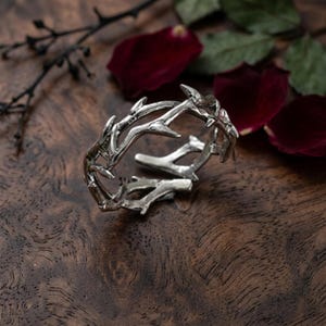 May include: A silver-toned ring designed to resemble a crown of thorns, featuring a textured, organic branch-like structure with sharp, protruding points. The metal has a polished finish that reflects light, creating a detailed, layered appearance.