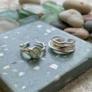 May include: Two silver rings. One ring features a textured, organic silver band holding a small, round, pale green stone. The second ring consists of multiple smooth, wavy silver bands layered together to create a stacked, open-ended design.