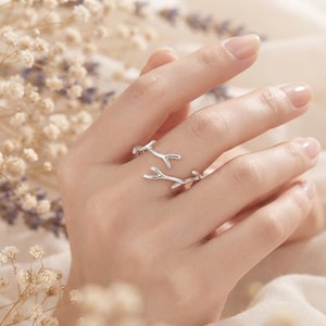 May include: A silver-toned open ring featuring a design of two delicate, branching twigs that overlap slightly. The metal has a smooth, polished finish, and the band is adjustable, showcasing a nature-inspired aesthetic with slender, curved ends.