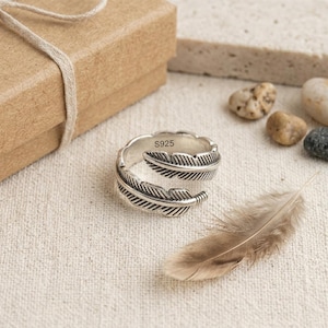 May include: A silver-toned open ring shaped like a feather with detailed etched lines. The inner band of the ring is clearly stamped with the text S925, indicating its composition. The ring has a wrap-around design with overlapping ends.