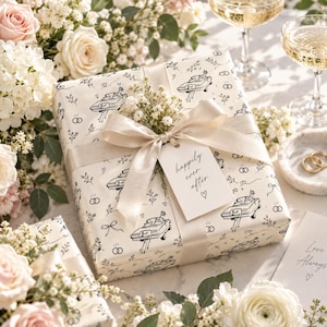 Könnte beinhalten: Ein quadratisches Geschenkpaket in cremefarbenem Papier mit schwarzem Linienmuster aus Autos, Eheringen und Schleifen. Ein beiges Satinband ist um das Paket gebunden und hält ein weißes Geschenketikett mit der Aufschrift "happily ever after".