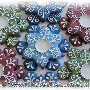 May include: Six snowflake-shaped gingerbread cookies decorated with white icing and a teal, green, blue, and burgundy glaze. Each cookie has a small, round indentation in the center to hold a tea light.