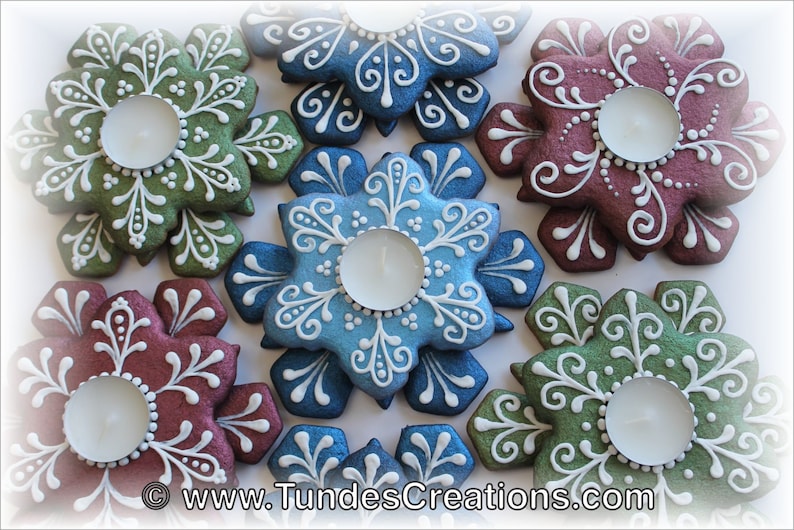 May include: Six snowflake-shaped gingerbread cookies decorated with white icing and a teal, green, blue, and burgundy glaze. Each cookie has a small, round indentation in the center to hold a tea light.