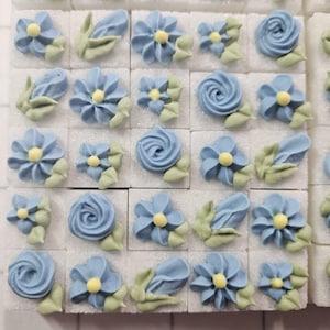 May include: A collection of 25 blue and yellow sugar flower decorations arranged in a grid pattern on a white surface. The flowers are made of buttercream frosting and are perfect for decorating cakes and cupcakes.