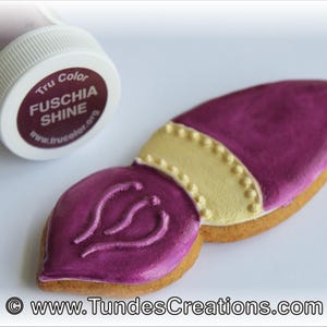 May include: A pink and gold paintbrush cookie decorated with a gold trim and a pink swirl design. The cookie is next to a small jar of fuchsia shine edible food coloring.