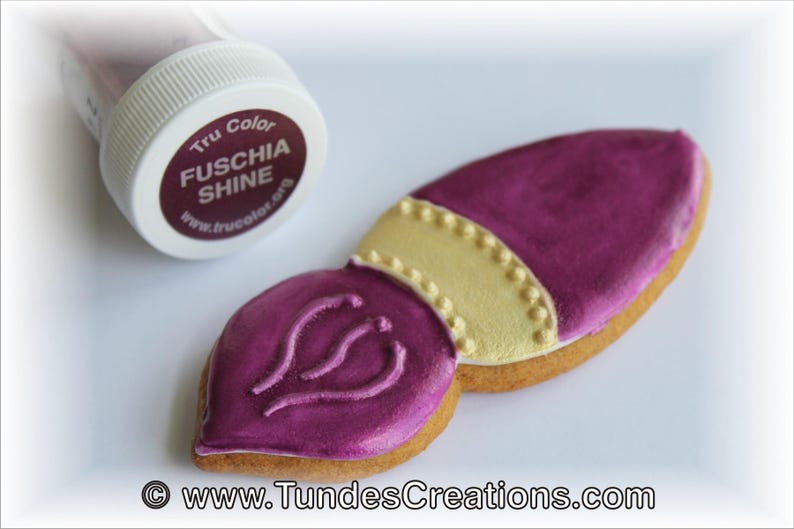 May include: A pink and gold paintbrush cookie decorated with a gold trim and a pink swirl design. The cookie is next to a small jar of fuchsia shine edible food coloring.