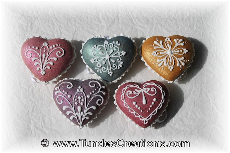 May include: Five heart-shaped cookies decorated with white icing and intricate designs. The cookies are in shades of pink, purple, teal, and gold.