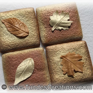 May include: Four square tiles with a textured surface in shades of gold and brown. Each tile features a different leaf design made of polymer clay, with colors ranging from gold to brown.