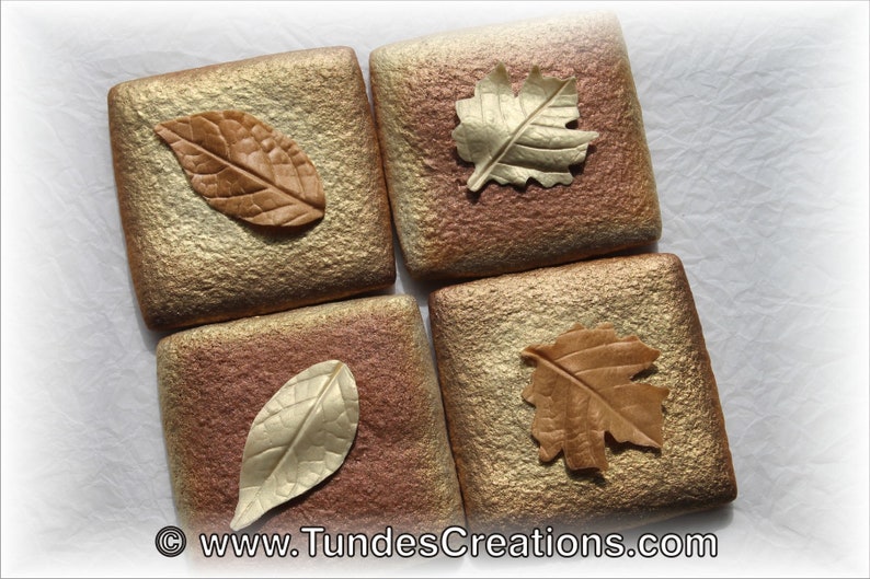 May include: Four square tiles with a textured surface in shades of gold and brown. Each tile features a different leaf design made of polymer clay, with colors ranging from gold to brown.