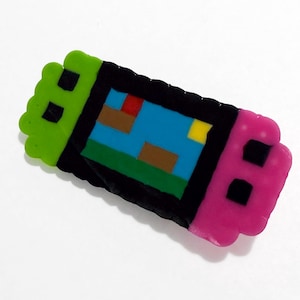 May include: A rectangular pixel art charm made of fused plastic beads. The design features a bright green left side, a pink right side, and a central black border surrounding a blue screen with small brown, red, and yellow pixelated shapes.