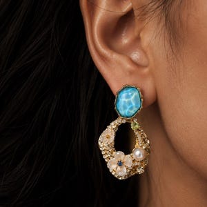 Puede incluir: Un pendiente colgante de tono dorado con una piedra azul ovalada en la parte superior de superficie ondulada. La sección inferior es un aro dorado abierto y texturizado adornado con dos flores blancas, una perla y cristales claros.
