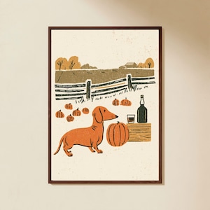 May include: A framed art print featuring a tan dachshund standing in a field with orange pumpkins, a wooden crate, a glass, and a bottle. The background shows a fence, trees, and a building under a cream-coloured sky with a textured, speckled finish.