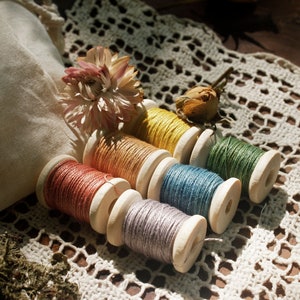May include: Six spools of colorful embroidery floss wound on wooden bobbins. The floss colors are orange, yellow, green, blue, purple, and brown. The spools are arranged on a white crocheted doily.